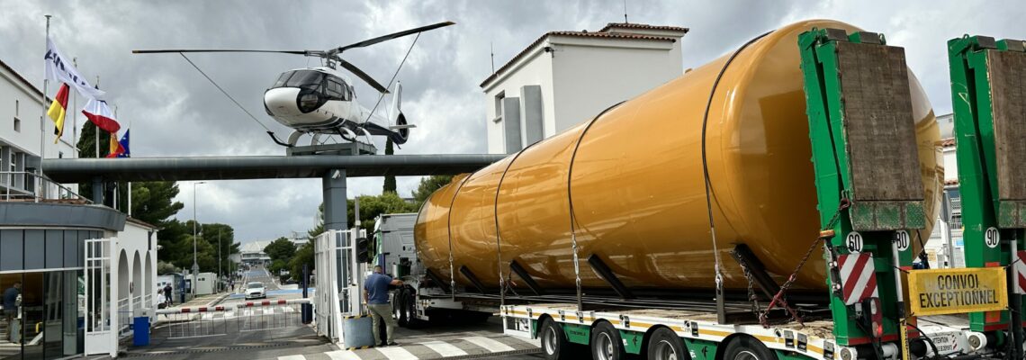 Transport exceptionnel d’une grande cuve cylindrique sur remorque, entrant dans un site industriel sous un hélicoptère statique.