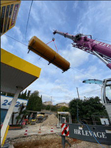 Levage d’une cuve cylindrique par une grue sur un chantier de station-service, avec zone de travaux et engins de chantier visibles.